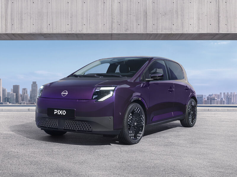 Three-quarter front view of a purple Nissan PIXO electric city car parked beneath a modern concrete structure, with a city skyline in the background.